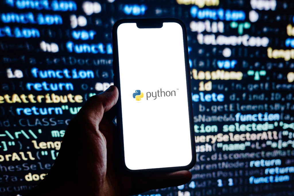 Auf einem Smartphone ist das Python-Logo zu erkennen. Das Smartphone wird von einer Hand vor einem Monitor gehalten, auf dem Python Codezeilen zu sehen sind.